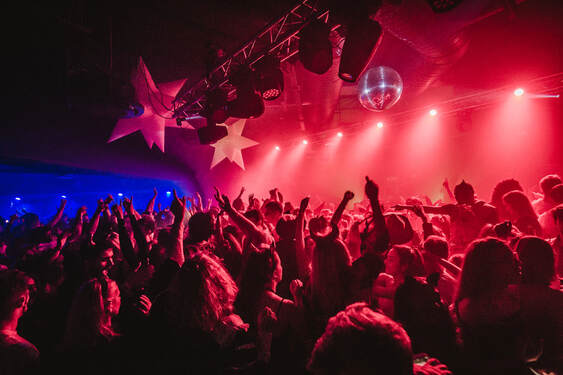 A busy nightclub filled with young people dancing with their arms in the air. The stage to the right is lit with six pink floodlights and a disco ball. The bar in the background is lit with electric blue spotlights, contrasting the otherwise warm rose-coloured lighting.